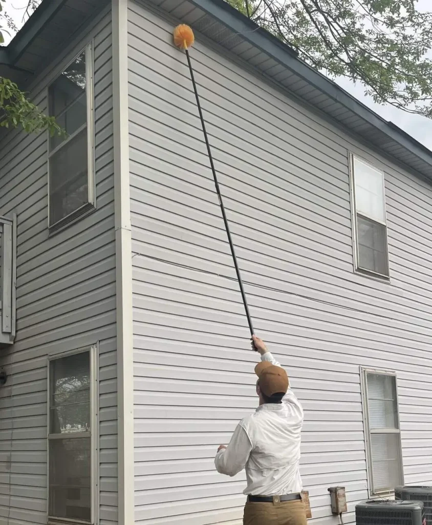 Wasp nest removal service in Huntsville AL. The Technician is removing a wasp nest with a pole on the side of a house.